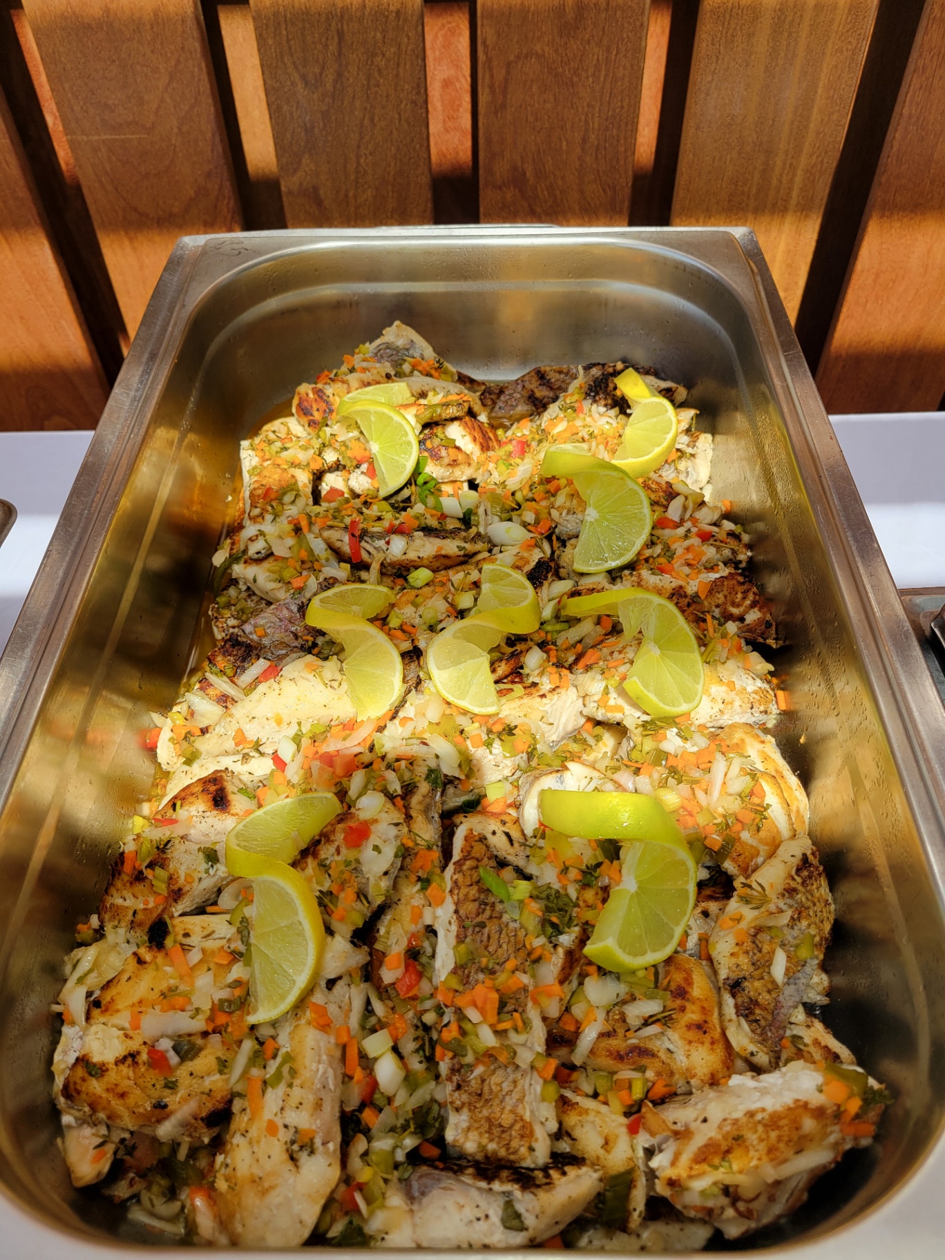 Chafing tray of grilled fish fillets topped with diced peppers, herbs and lime wedges